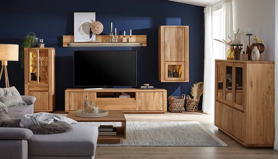 Gemütliches Wohnzimmer mit dunkler Akzentwand, massiven Eichenmöbeln – Vitrine, TV-Board und Sideboard – grauem Ecksofa, flauschigem Teppich und großer Fensterfront  